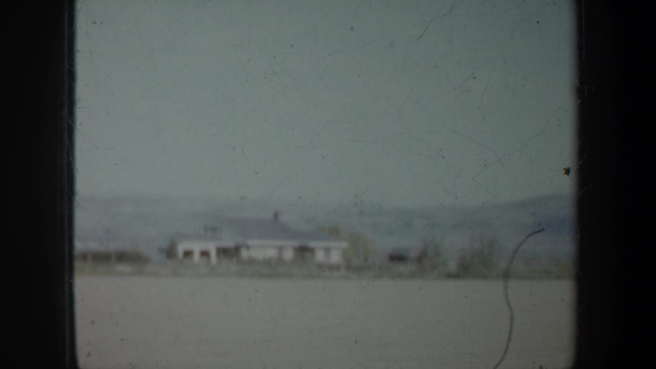1957 Oregon home-movie frame showing an open rural field, distant white building, tree line, and low hills from a moving vehicle.