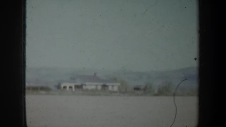 1957 Oregon home-movie frame showing an open rural field, distant white building, tree line, and low hills from a moving vehicle.