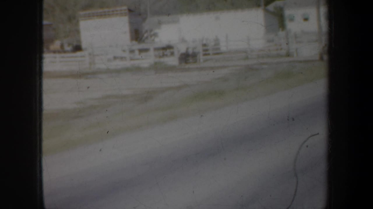 Moving-vehicle view of rural buildings, white fencing, and a roadside area in the Portland, Oregon 1957 home-movie clip.