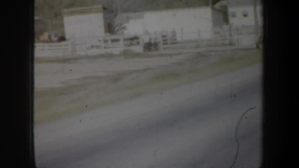 Moving-vehicle view of rural buildings, white fencing, and a roadside area in the Portland, Oregon 1957 home-movie clip.