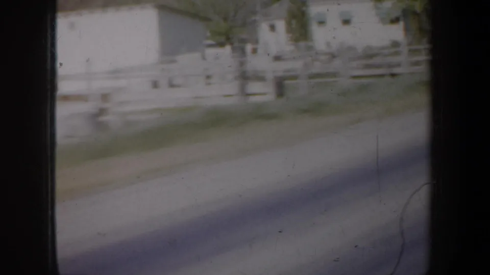 1957 Oregon frame with motion-blurred white fencing, roadside surface, and pale rural buildings viewed from a moving vehicle.