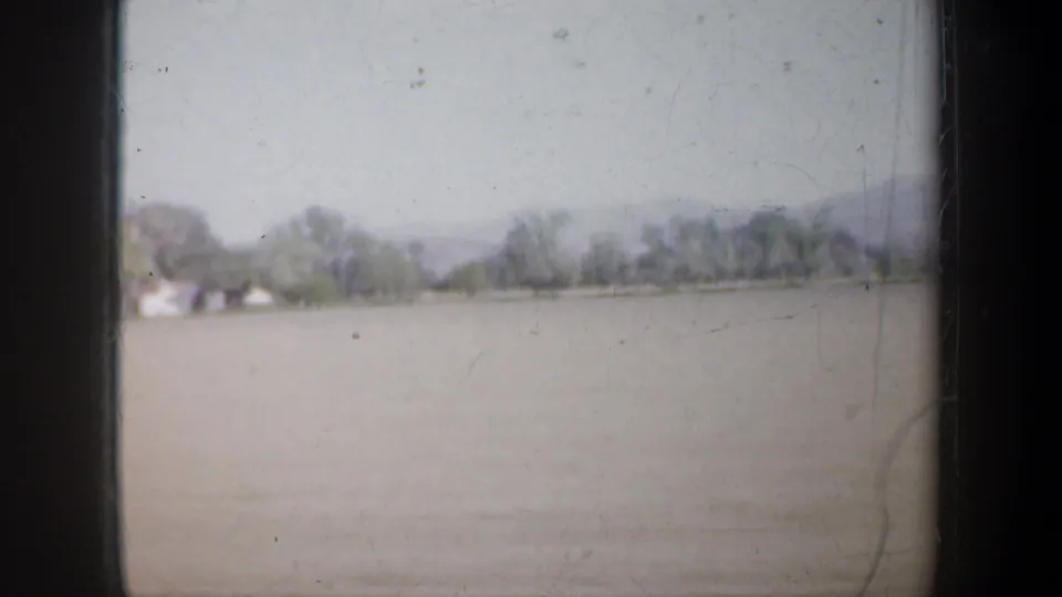 Wide rural field near Portland, Oregon, with distant trees and small buildings in a soft 1957 8mm home-movie frame.