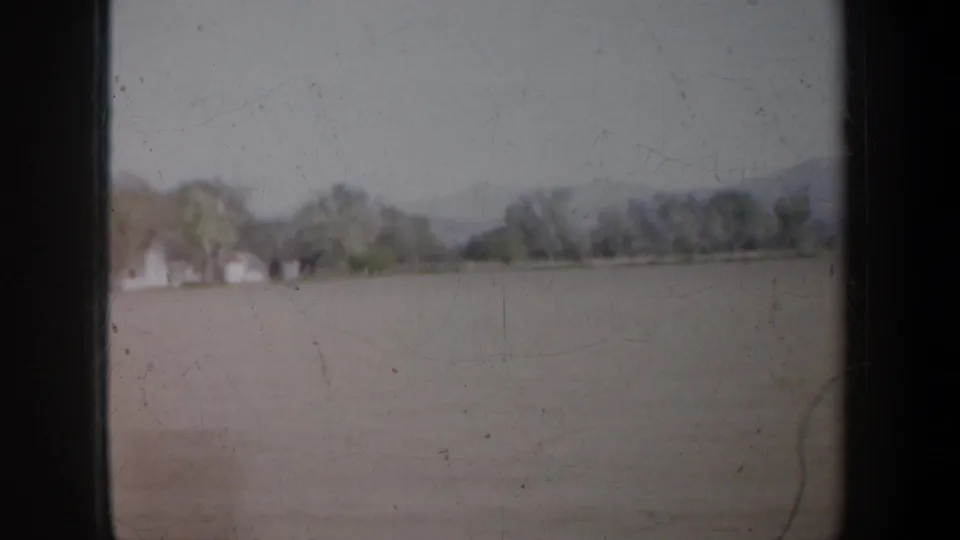 1957 Portland-area 8mm view from a moving vehicle, showing a pale field, distant houses, trees, and a soft horizon.