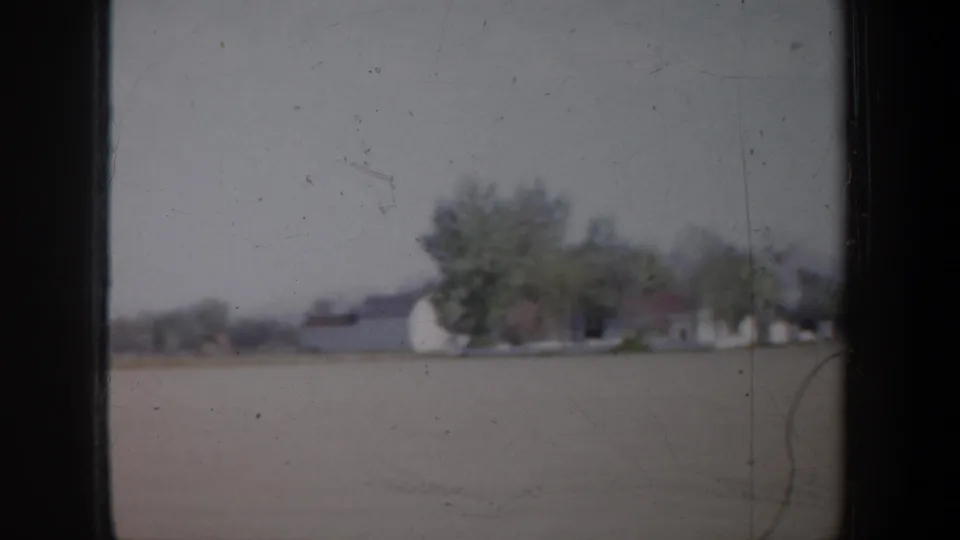 Soft 1957 8mm roadside frame showing a low house, clustered trees, and a broad field in the Portland area.