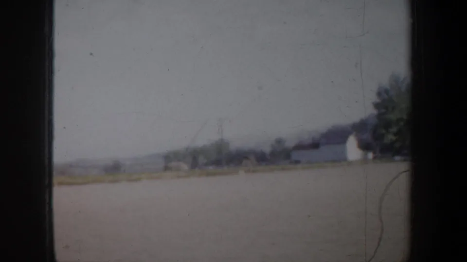 Portland-area 1957 8mm driving view with a distant building, trees, open field, and faint low hills on the horizon.
