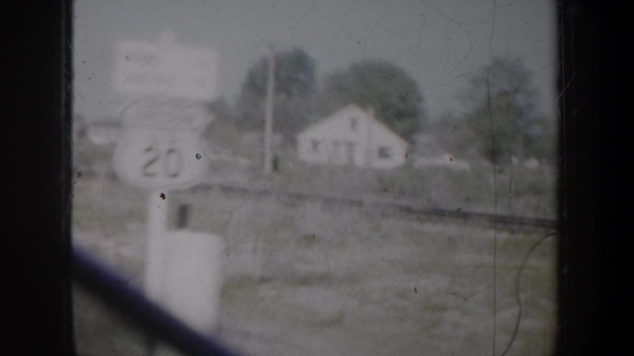 1957 Portland-area home-movie frame showing a blurred rural house, roadside marker, and dark window framing from a moving vehicle.