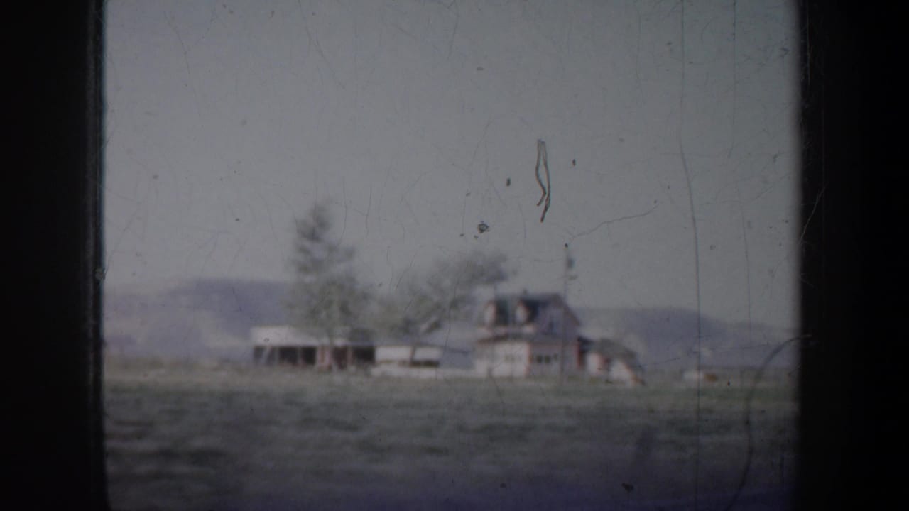 Distant rural house, open field, trees, and low hills seen past a window divider.
