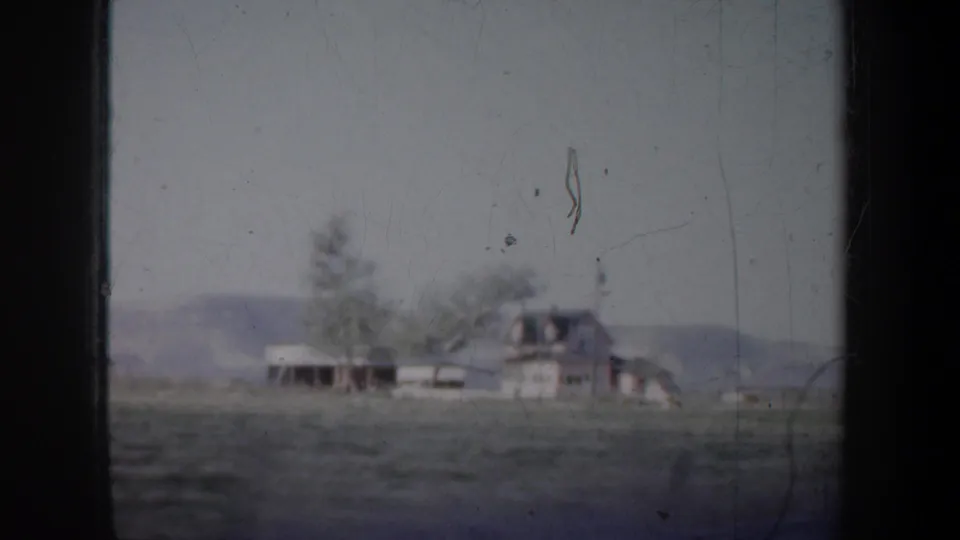 1957 Portland-area 8mm frame with a distant house, open field, trees, and low hills viewed through a vehicle window.