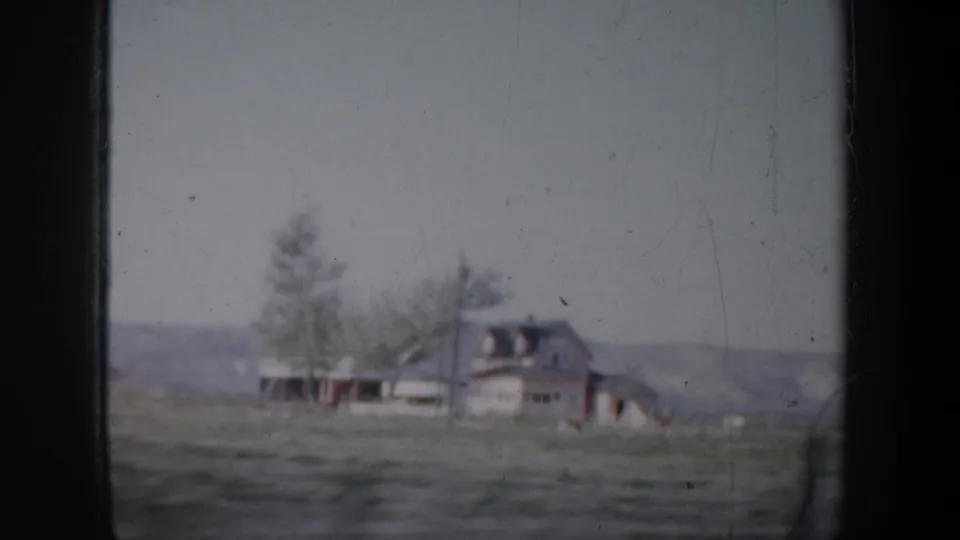Soft 1957 Portland-area home-movie frame showing a rural house, open ground, trees, and distant hills through a vehicle window.