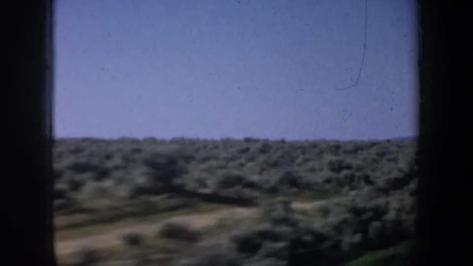 1957 Portland-area travel frame with blurred green fields, a pale road or track, low horizon, and blue sky from a moving vehicle.