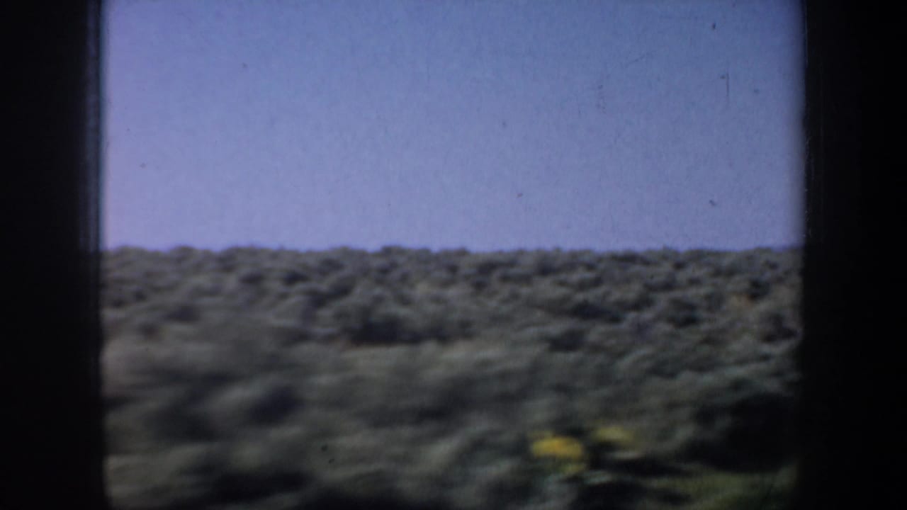1957 8mm home-movie view of open western scrubland through a dark window frame, with a hazy sky and motion softness.
