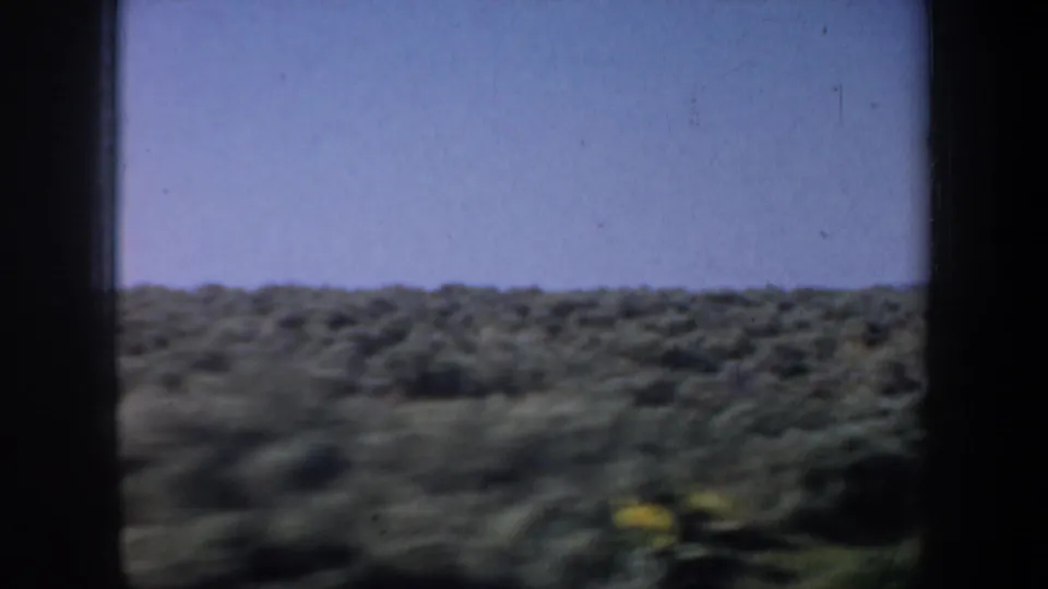 1957 8mm home-movie view of open western scrubland through a dark window frame, with a hazy sky and motion softness.