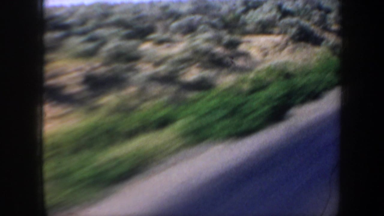 A moving 1957 8mm vehicle view shows a paved roadside edge, green shrubbery, dusty ground, and blurred western terrain.
