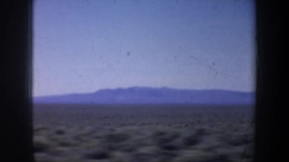 1957 8mm home-movie frame with distant low mountains, open scrubland, and a pale blue sky seen from a moving vehicle.