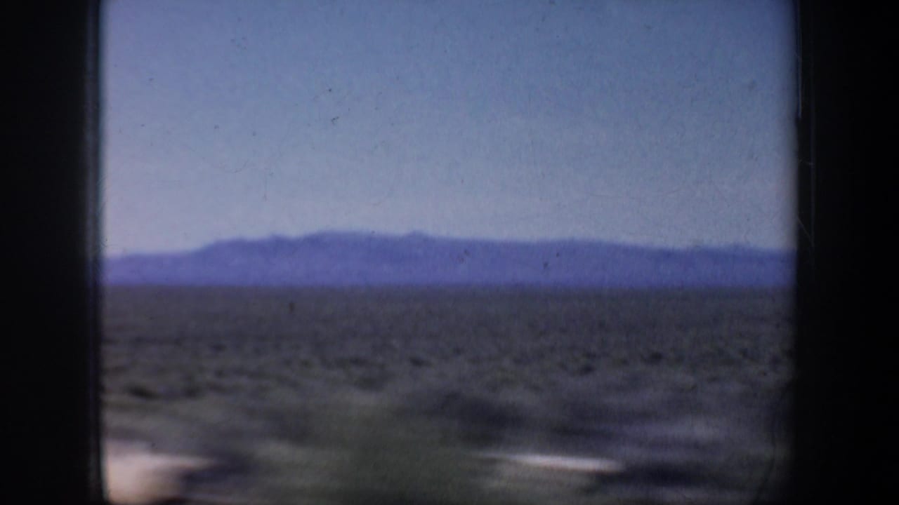 Distant mountain ridge and scrubland pass behind a dark vertical vehicle-window frame.