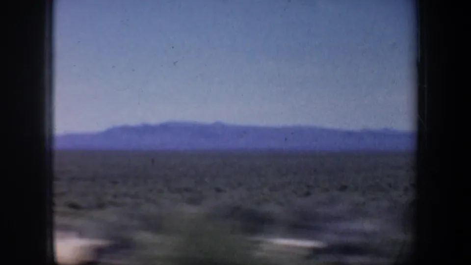 Arid terrain and a purple-toned mountain ridge appear beyond a vehicle-window frame in 1957 home-movie footage.