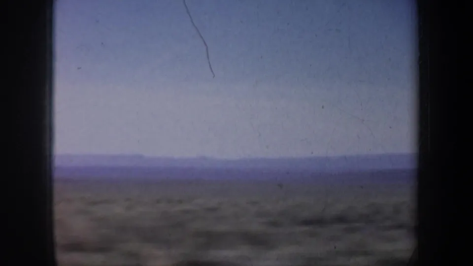 A soft 8mm view of open arid ground and distant low mountains, with scratches and motion blur from vehicle travel.