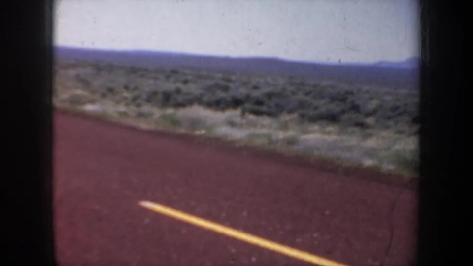 1957 8mm travel frame showing a red road surface and yellow center line passing arid scrubland and distant mountains.