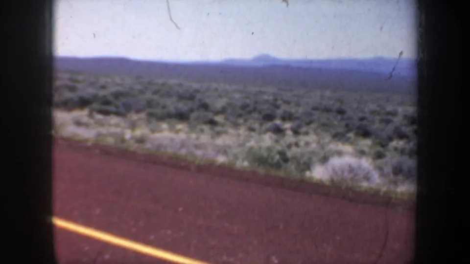 1957 8mm home-movie road view across desert scrub, with a yellow center line and low mountain horizon.