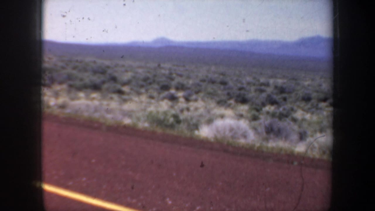 1957 8mm road-trip view from a moving vehicle, showing arid scrubland, distant mountains, pale sky, and the edge of a red road surface.