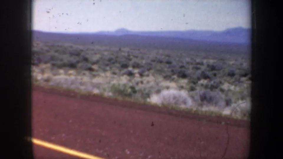 1957 8mm road-trip view from a moving vehicle, showing arid scrubland, distant mountains, pale sky, and the edge of a red road surface.