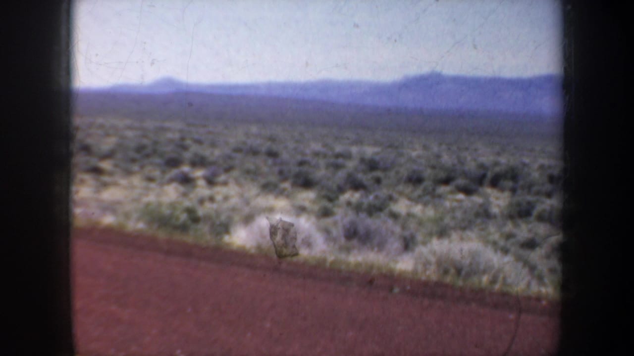 Open scrubland and distant mountain ridge seen beyond a red road edge through a dark-framed view.