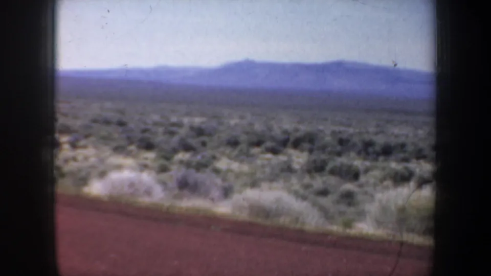 1957 8mm vehicle-window view of desert shrubs, roadside grasses, distant mountains, and a red road surface softened by motion.
