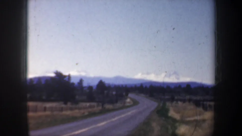 1957 8mm road-trip view shows a curving paved road, roadside fields, dark trees, and distant hills or mountains.