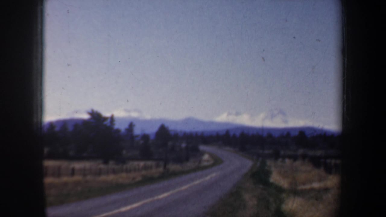 1957 Oregon 8mm view from a vehicle window of a rural road, fields, tree line, and distant snow-capped mountains.