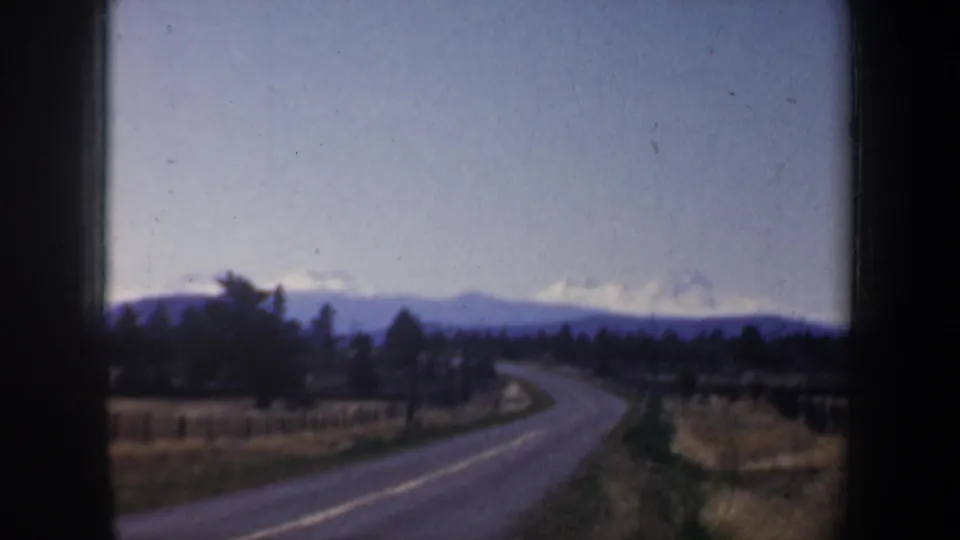 1957 Oregon 8mm view from a vehicle window of a rural road, fields, tree line, and distant snow-capped mountains.
