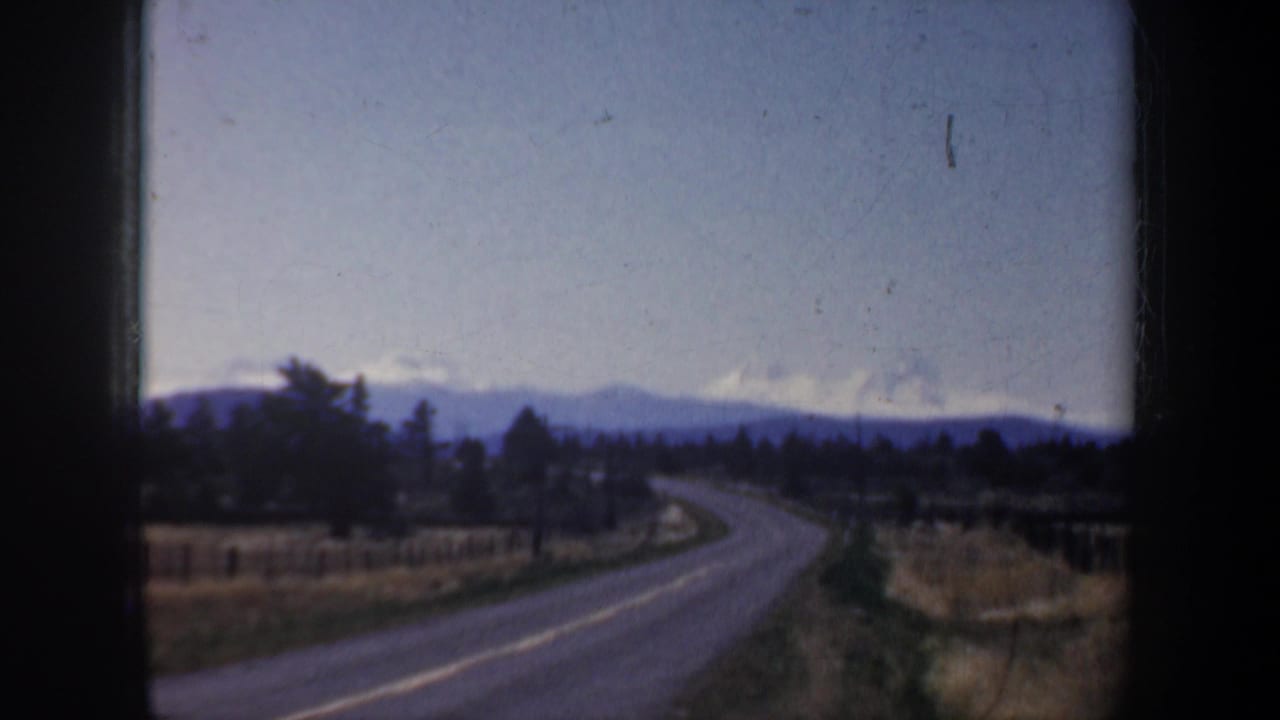 Roadside countryside with a curving road, trees, fields, and distant mountain ridge.