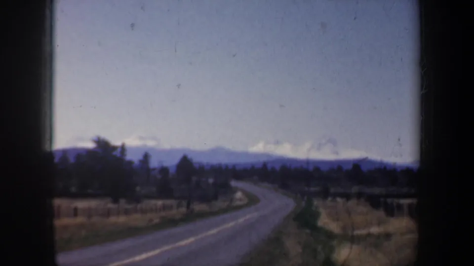 Soft-focused 1957 Oregon road view with fields, trees, distant hills, and snow-capped mountains seen through a vehicle window.