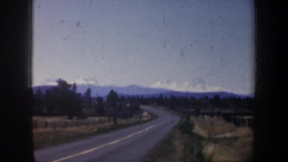 1957 Portland-area 8mm frame of a rural road and open fields, with evergreen trees and distant snow-capped mountains beyond.