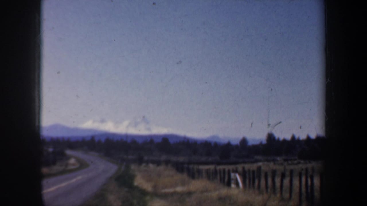 1957 Oregon home-movie frame showing a rural road curving past fences toward distant mountains near Portland.