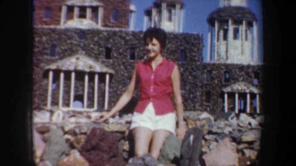 1957 Portland home-movie frame of a smiling woman posing near rocks before a detailed miniature classical-style building.