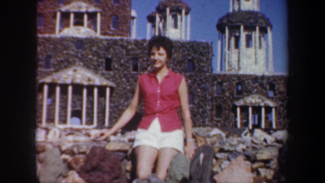 In 1957 Portland home-movie footage, a woman poses beside stone walls, columns, and small domed towers at a landmark attraction.