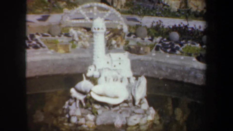 1957 Portland home-movie frame of a small lighthouse model set among shells, stones, and pond edging.