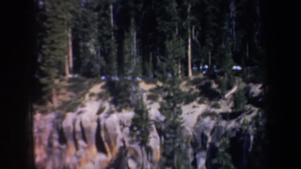 1957 Oregon home-movie frame looking across evergreen trees toward a grooved rocky cliff face.