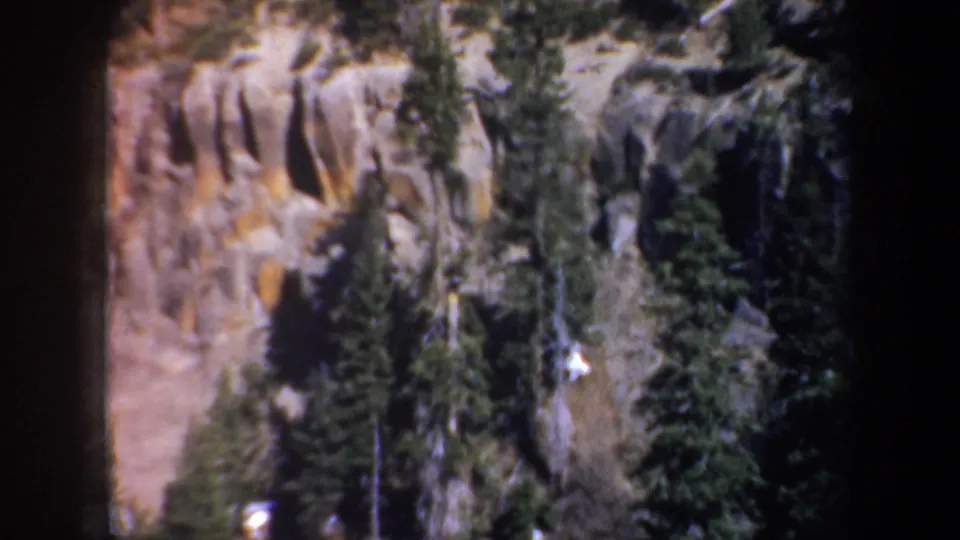1957 Oregon home-movie frame showing the lower rocky canyon face with evergreens and heavy edge darkening.