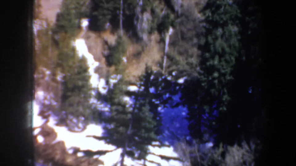 1957 home-movie frame of the lower portion of a rocky forest slope, with bright snow crossing the foreground area.