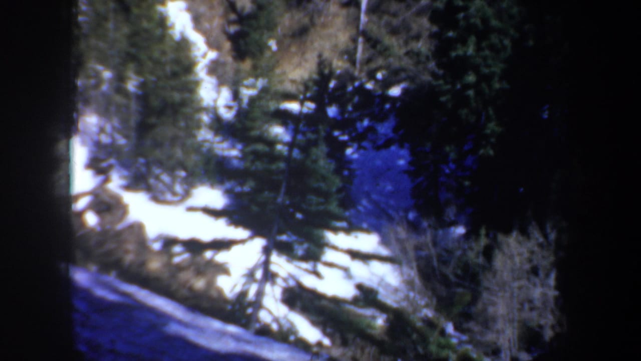1957 Oregon home-movie frame showing snow patches, evergreens, and rocky mountain terrain under cool blue light.