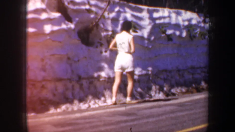Woman in a white sleeveless outfit walks away along a paved roadside beside a stone retaining wall in 1957 Portland, Oregon footage.