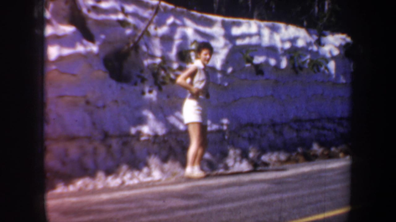 1957 home-movie frame of a young woman in white standing near a stone wall and roadside pavement in Oregon.