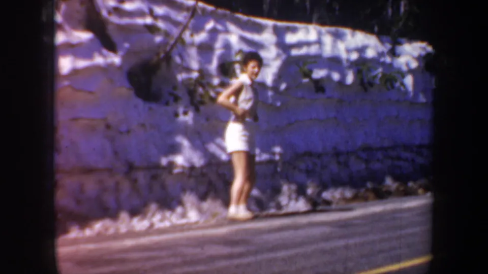1957 home-movie frame of a young woman in white standing near a stone wall and roadside pavement in Oregon.