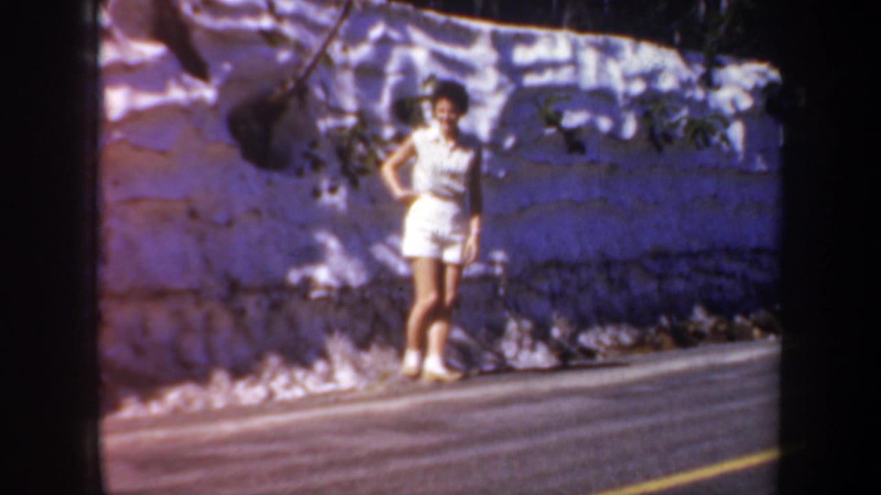 Woman standing near a roadside stone wall with dappled shadows.