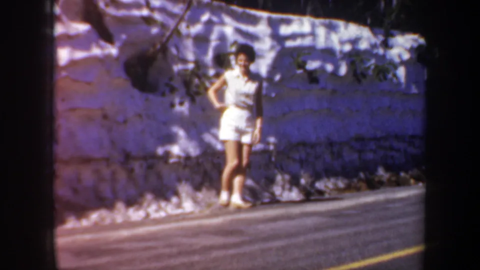 Young woman in white stands near the wall and road in a 1957 Oregon home-movie travel scene.