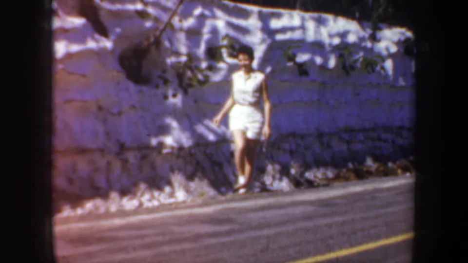 A 1957 8mm frame shows the woman moving along the roadside with the layered stone wall behind her.