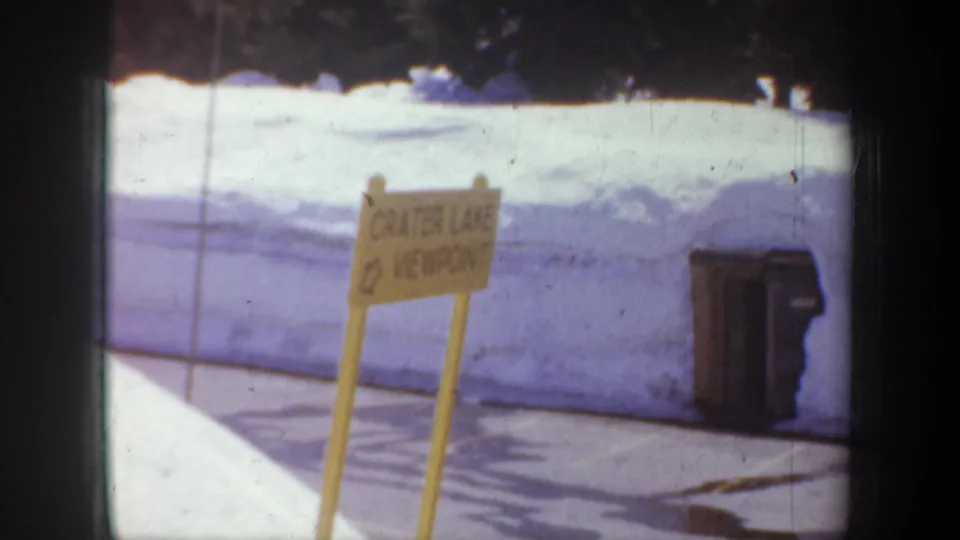 A yellow sign reading Crater Lake Viewpoint stands near roadside pavement and a pale wall in the 1957 Oregon footage.