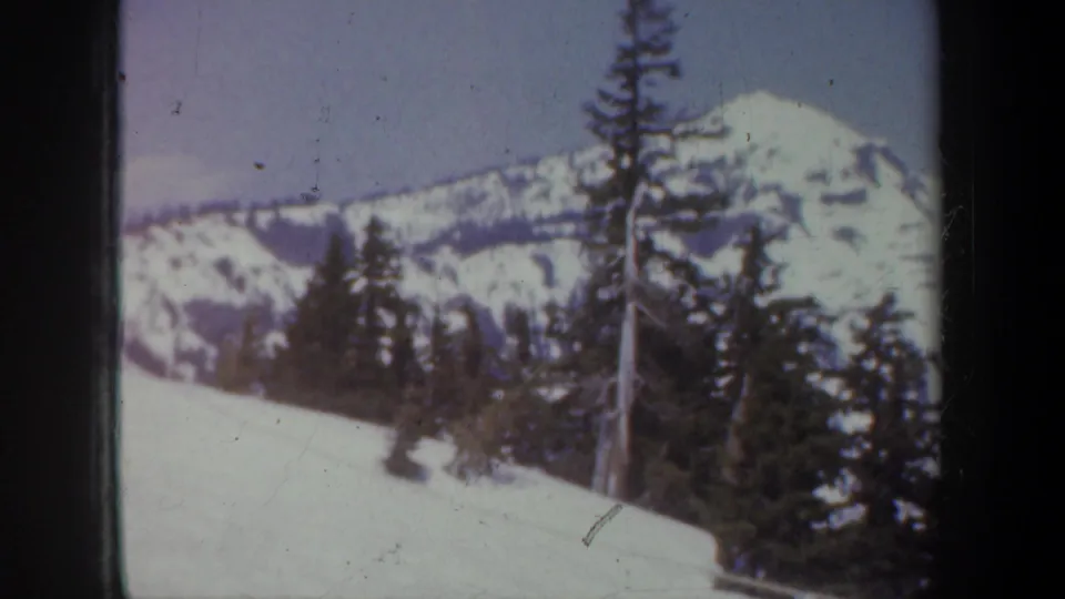 1957 Oregon home-movie frame showing snowy mountains, evergreen trees, and a white slope under a pale sky.