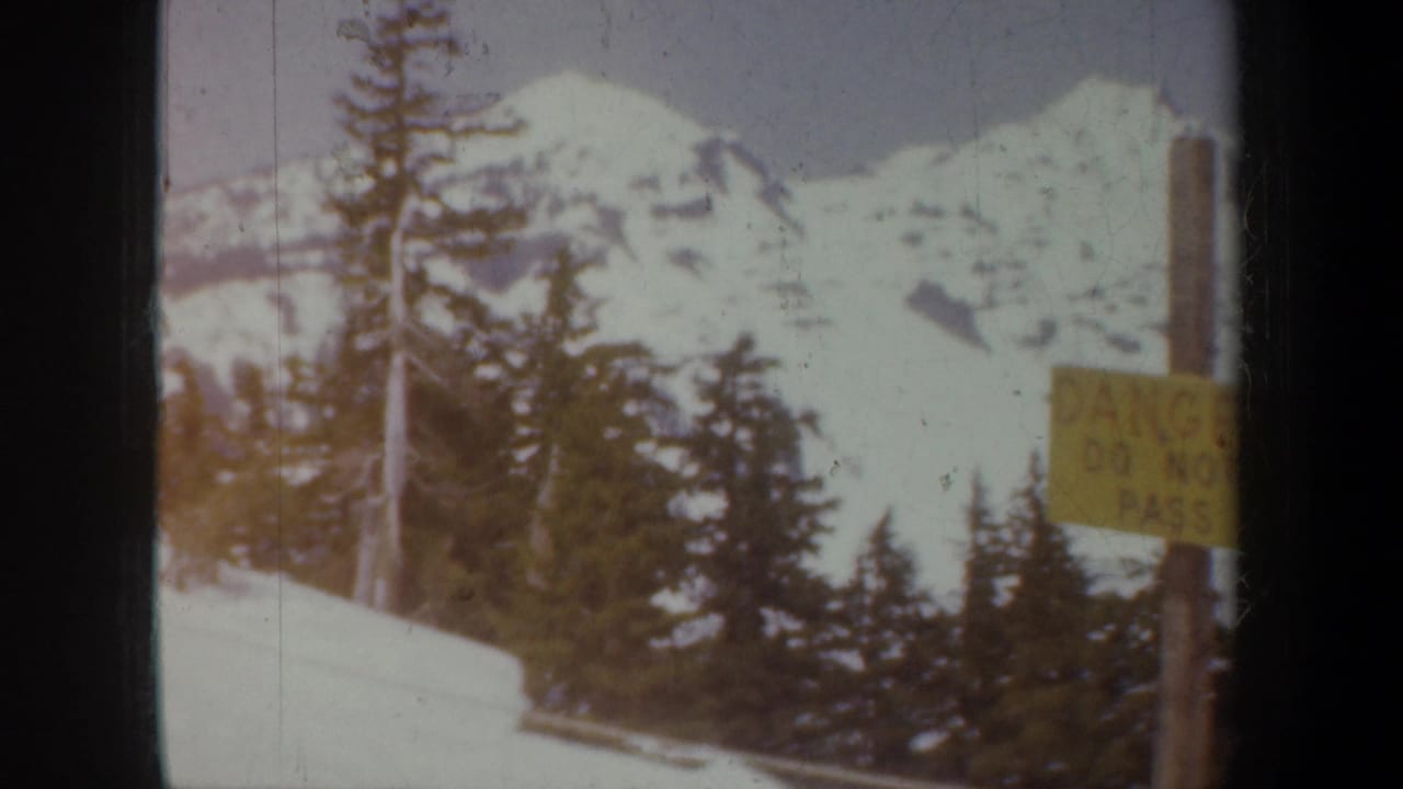 1957 Oregon home-movie frame showing snowy mountains, dark evergreen trees, and a yellow danger warning sign in a winter roadside view.